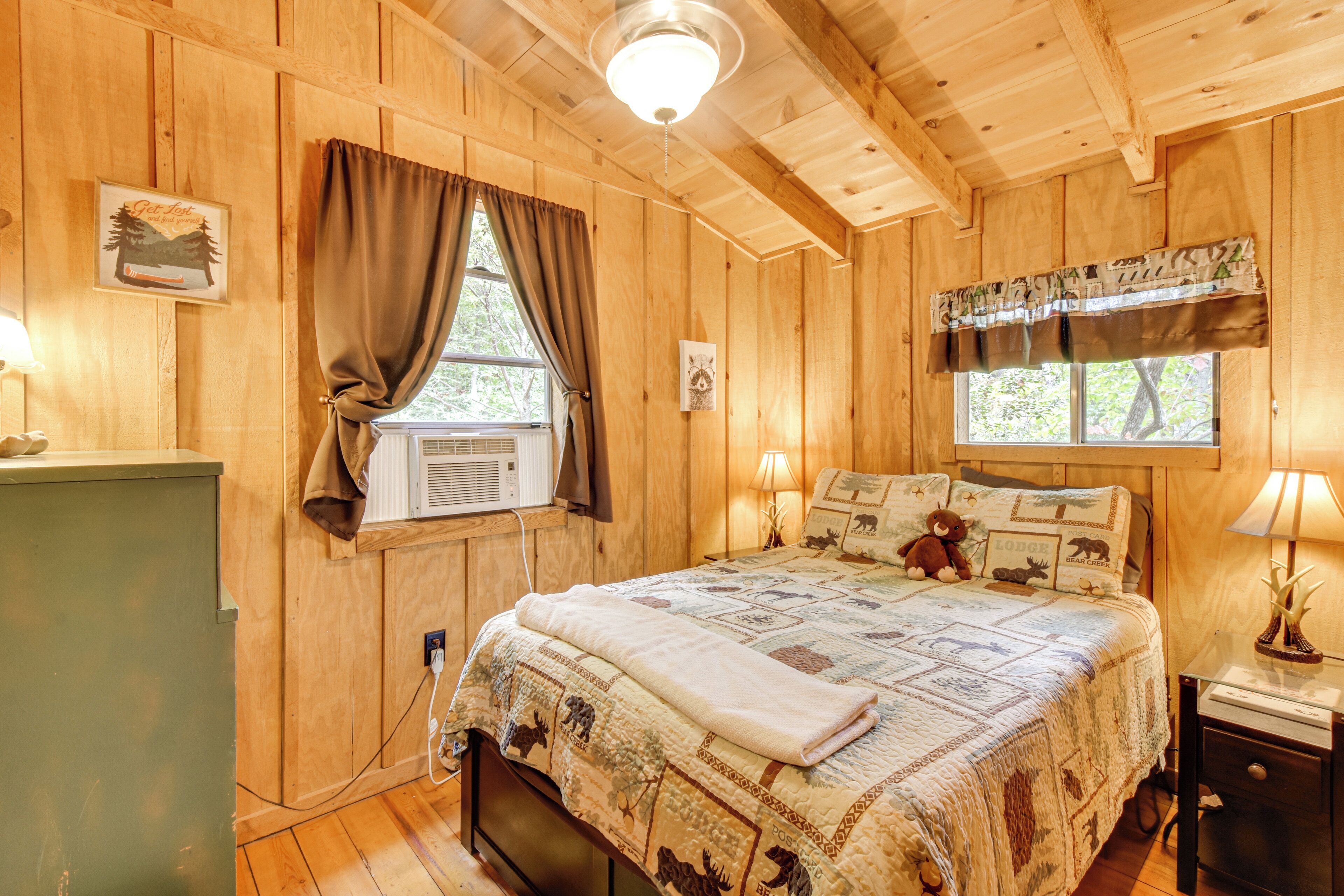 A wooden cabin bedroom with a bed, a nightstand, a lamp, a window with curtains, and a wall-mounted air conditioner.
