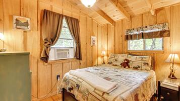 A wooden cabin bedroom with a bed, a nightstand, a lamp, a window with curtains, and a wall-mounted air conditioner.
