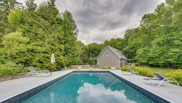 Una piscina rettangolare con acqua cristallina, circondata da una terrazza lastricata. Lungo il bordo della piscina ci sono lettini da relax. Si vede una casa con tetto a due falde sullo sfondo, immersa nel verde di alberi rigogliosi.