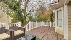 Une terrasse en bois avec une balustrade blanche, un canapé en osier agrémenté de coussins et une table avec un plateau en verre.
