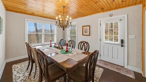 Une salle à manger avec un plafond en bois, un lustre, une table à manger dressée avec des sets de table et des bougies, et une porte dotée d’un store vénitien.