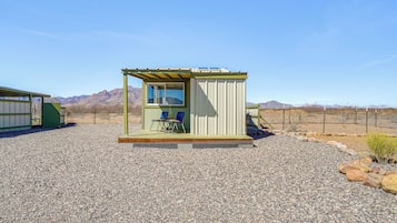 A small, single-room structure with a window and a door, a small porch with two chairs, and a gravel ground.