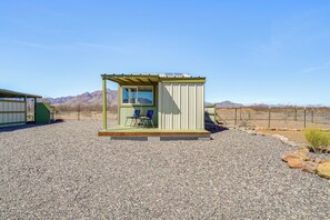 A small, single-room structure with a window and a door, a small porch with two chairs, and a gravel ground. - Tiny Town Cottages in Rodeo! 'dog House' (Rodeo)