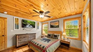 Un dormitorio con techo de madera, cama con colcha temática de oso, ventilador de techo y ventanas grandes con persianas.