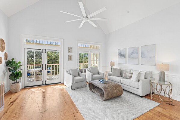A spacious living room with a white sofa, two armchairs, a coffee table, and a ceiling fan. Large windows provide natural light and a view of the outdoors. - Idle Hour in WaterColor | 2bd + Loft, 2 bikes, Steps to WaterColor Beach Club (Santa Rosa Beach)