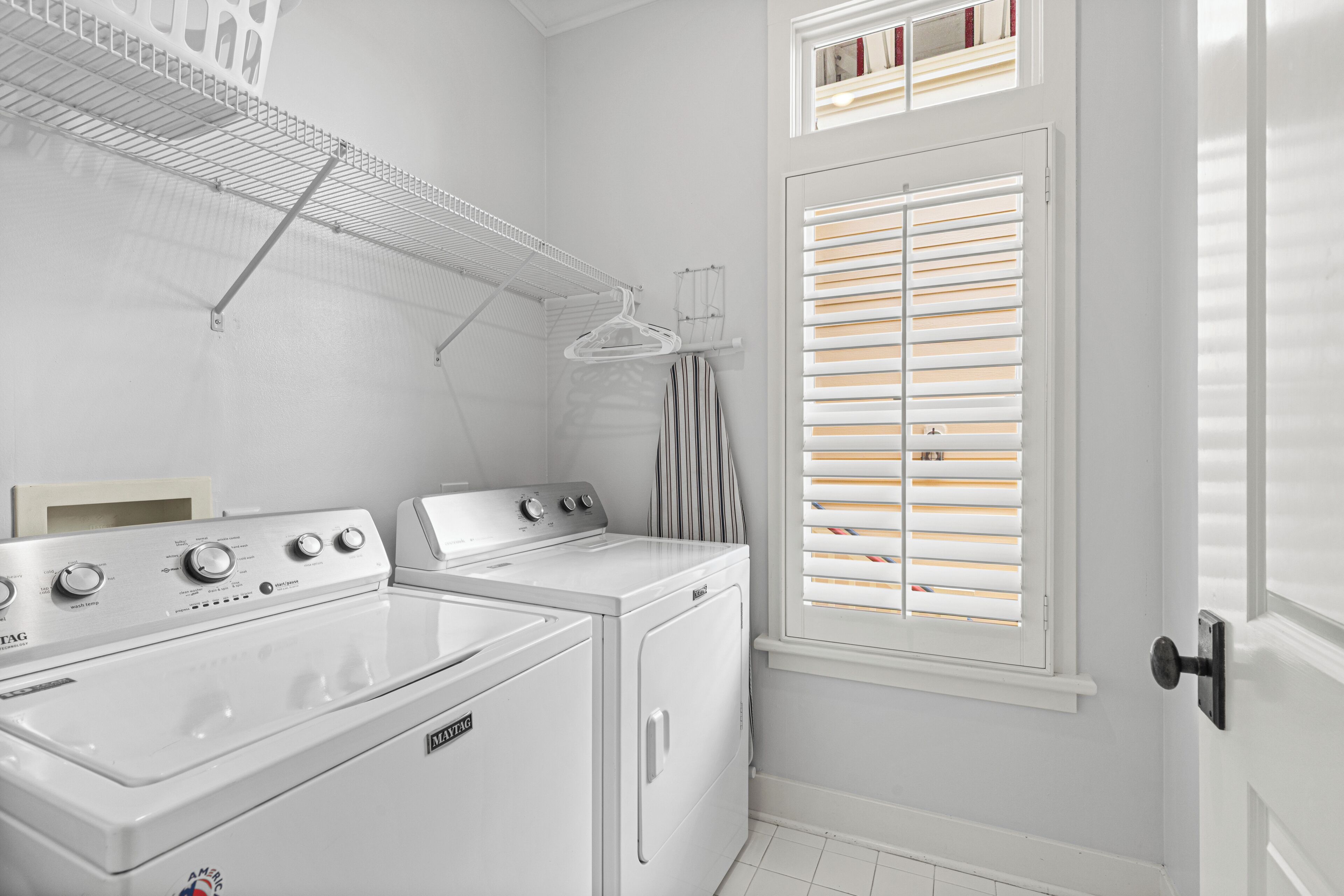 A laundry room with a front-loading washer and dryer, a drying rack, and a window with blinds.