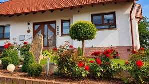 Una casa con techo de tejas, puerta principal y un pequeño jardín con flores rojas y un árbol esférico.
