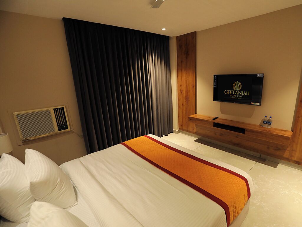 Ein Hotelzimmer mit Bett, Fernseher, einer Holzbank und einem großen Fenster mit Vorhängen.