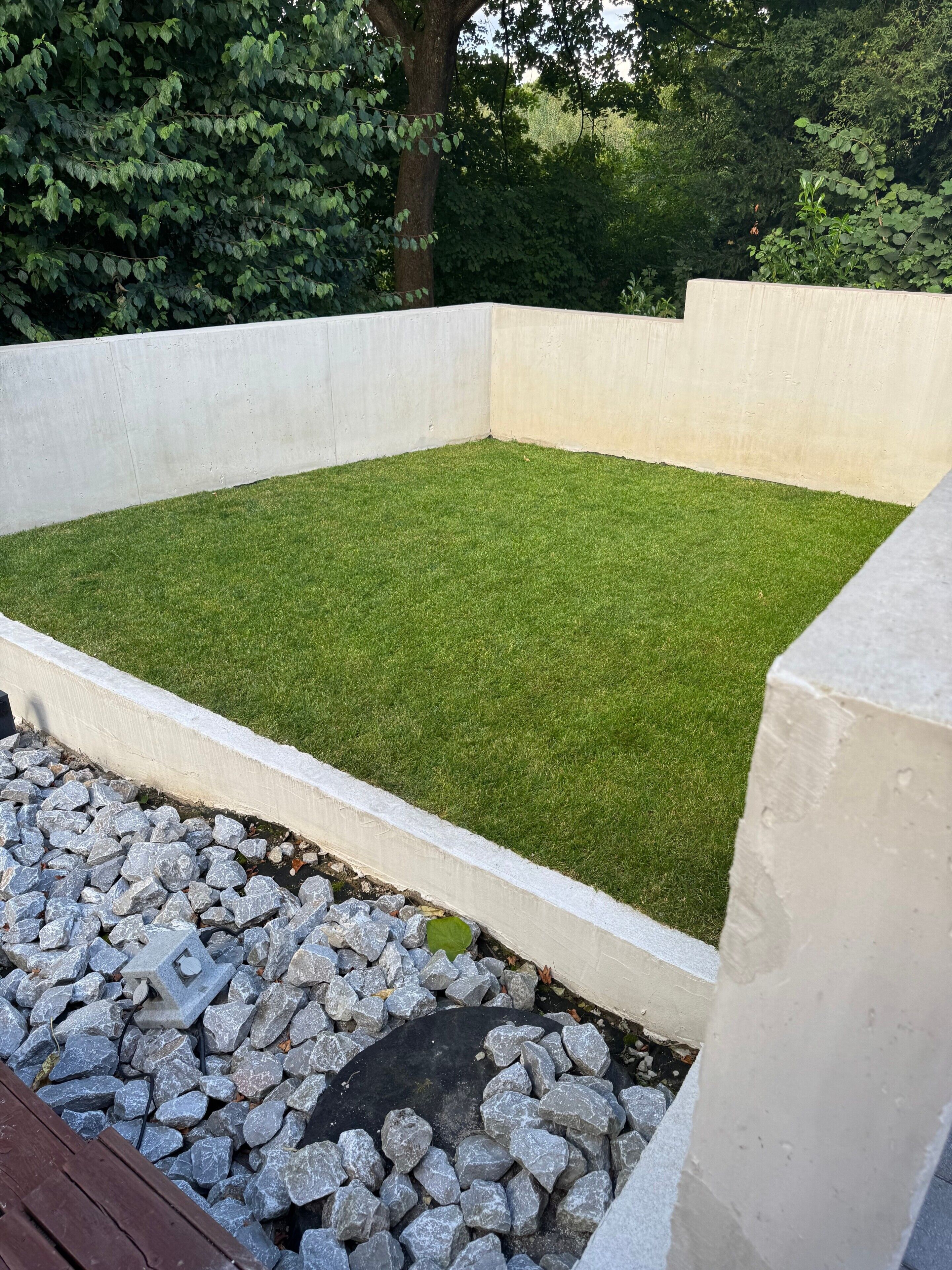 Zona de jardí rectangular amb superfície plana, envoltada per una paret baixa de formigó i un adorn de pedres a la part inferior.