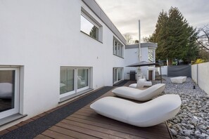 A modern outdoor deck with white lounge chairs, a wooden deck, and a white building with large windows. - Design Apartment with Whirlpool | Large Terrace by Park | Near University (Paderborn)