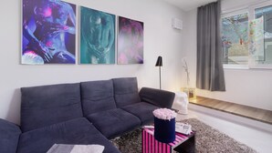 Un salon moderne avec un canapé gris foncé, des œuvres d’art abstraites accrochées au mur, une sculpture en buste blanche et une fenêtre avec des rideaux.