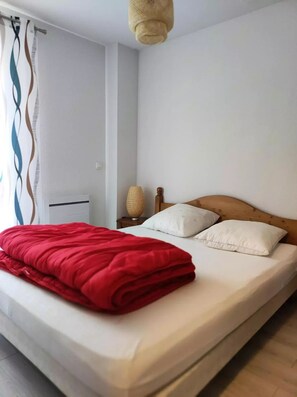 Un lit bien fait, avec une couverture rouge et deux oreillers, une lampe de chevet et une fenêtre avec un rideau.