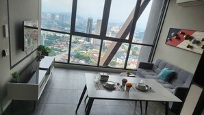 A modern room with a large window offering a city view, a dining table set with plates and cutlery, a grey sofa with a teal pillow, and a small potted plant on the windowsill. - Scarletz Suites klcc by Signature Apartment (Kuala Lumpur)