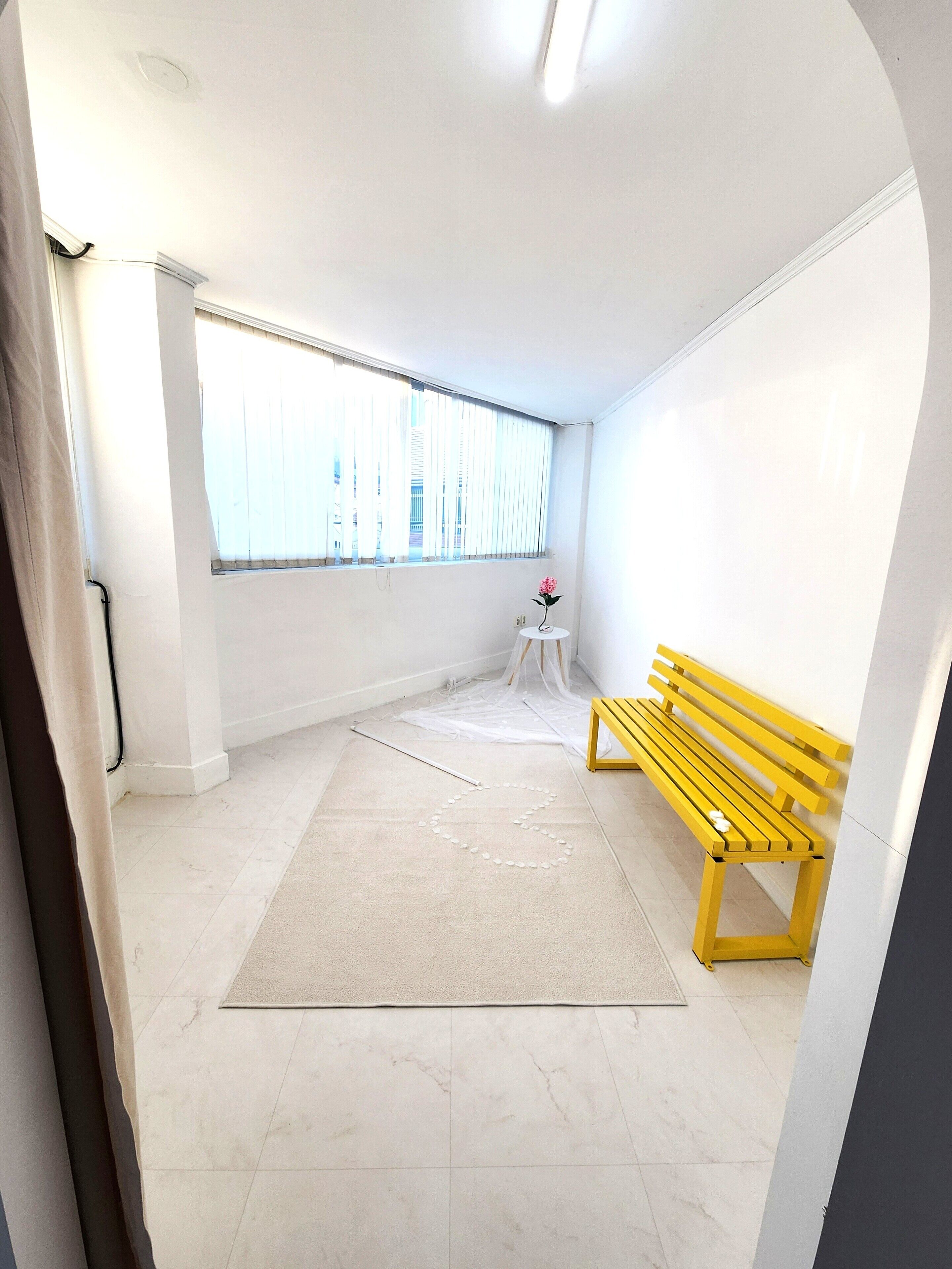 A room with a large window, a yellow bench, a small table with a flower, and a white floor.