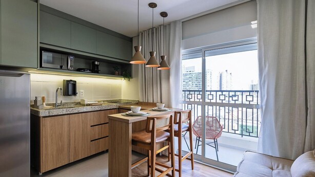 A modern kitchen with wooden cabinets, a built-in oven, and a coffee machine. There is a dining area with a table, chairs, and a view of the city through large windows. - Complete Studio 350 m From the Subway Ipi0034 (São Paulo)
