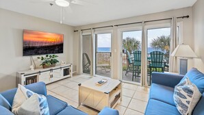 A living room with a sofa, coffee table, and a TV mounted on the wall. There is a balcony with chairs and a view of the beach. - Windancer 208 Gulf Front, Free Beach Service, Emerald Waters, Pool, & Grill. (Miramar Beach)
