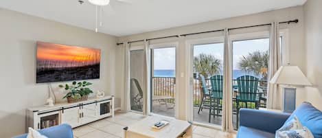Een woonkamer met een bank, salontafel en een aan de muur bevestigde televisie. Er is een balkon met stoelen en uitzicht op het strand.