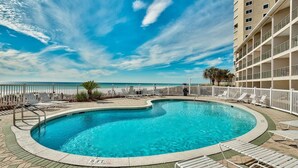 A pool with a curved edge, surrounded by lounge chairs and a fence, with a building in the background. - Windancer 208 Gulf Front, Free Beach Service, Emerald Waters, Pool, & Grill. (Miramar Beach)
