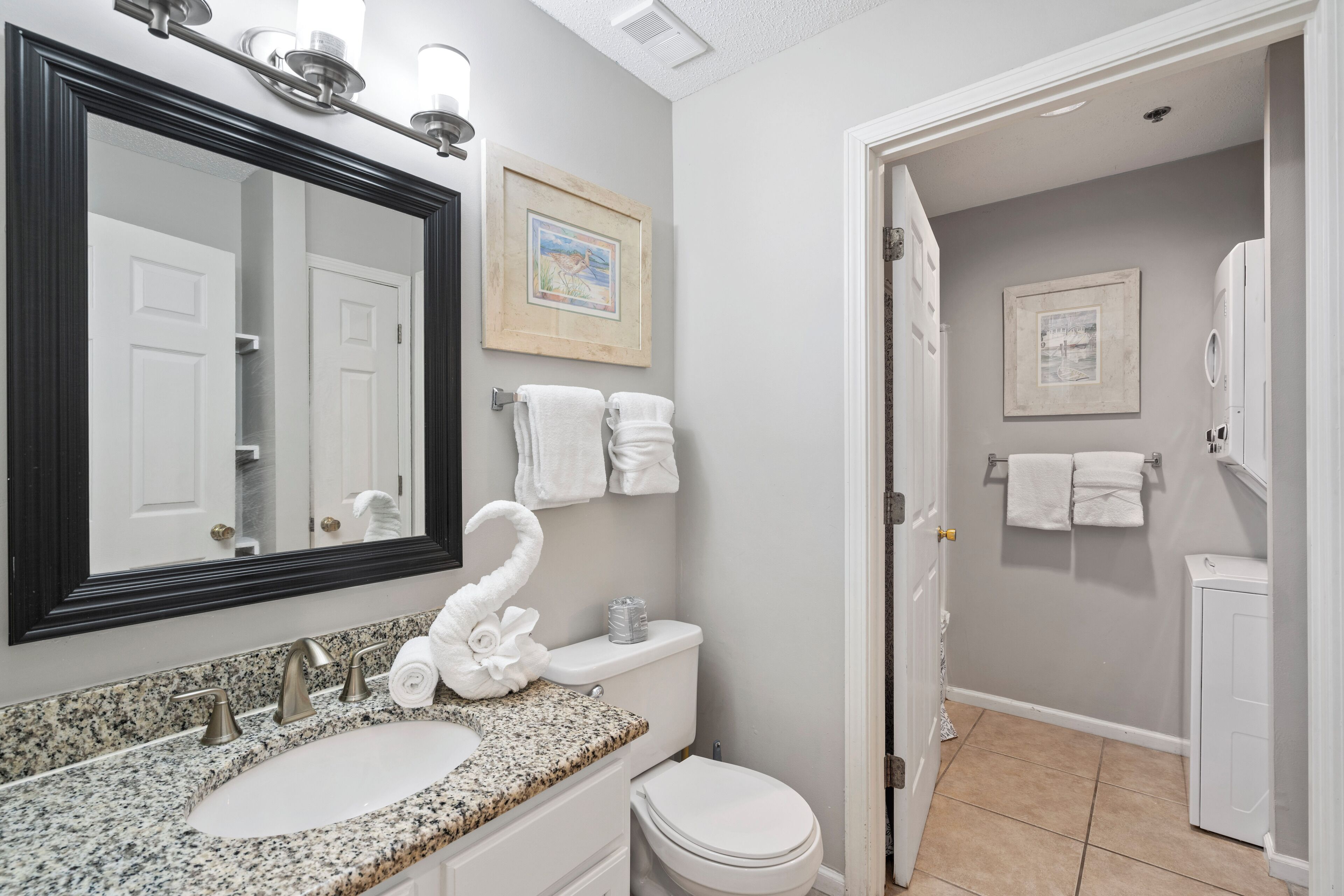 Une salle de bain avec un lavabo à vasque en granit, des toilettes blanches et un grand miroir avec un cadre noir. Deux tableaux encadrés ornent le mur, et un porte-serviettes contient des serviettes blanches.
