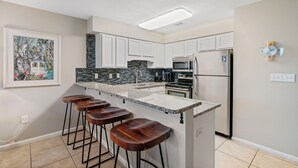 A modern kitchen with white cabinets, a granite countertop, a stainless steel refrigerator, and a bar stool area. - Windancer 208 Gulf Front, Free Beach Service, Emerald Waters, Pool, & Grill. (Miramar Beach)