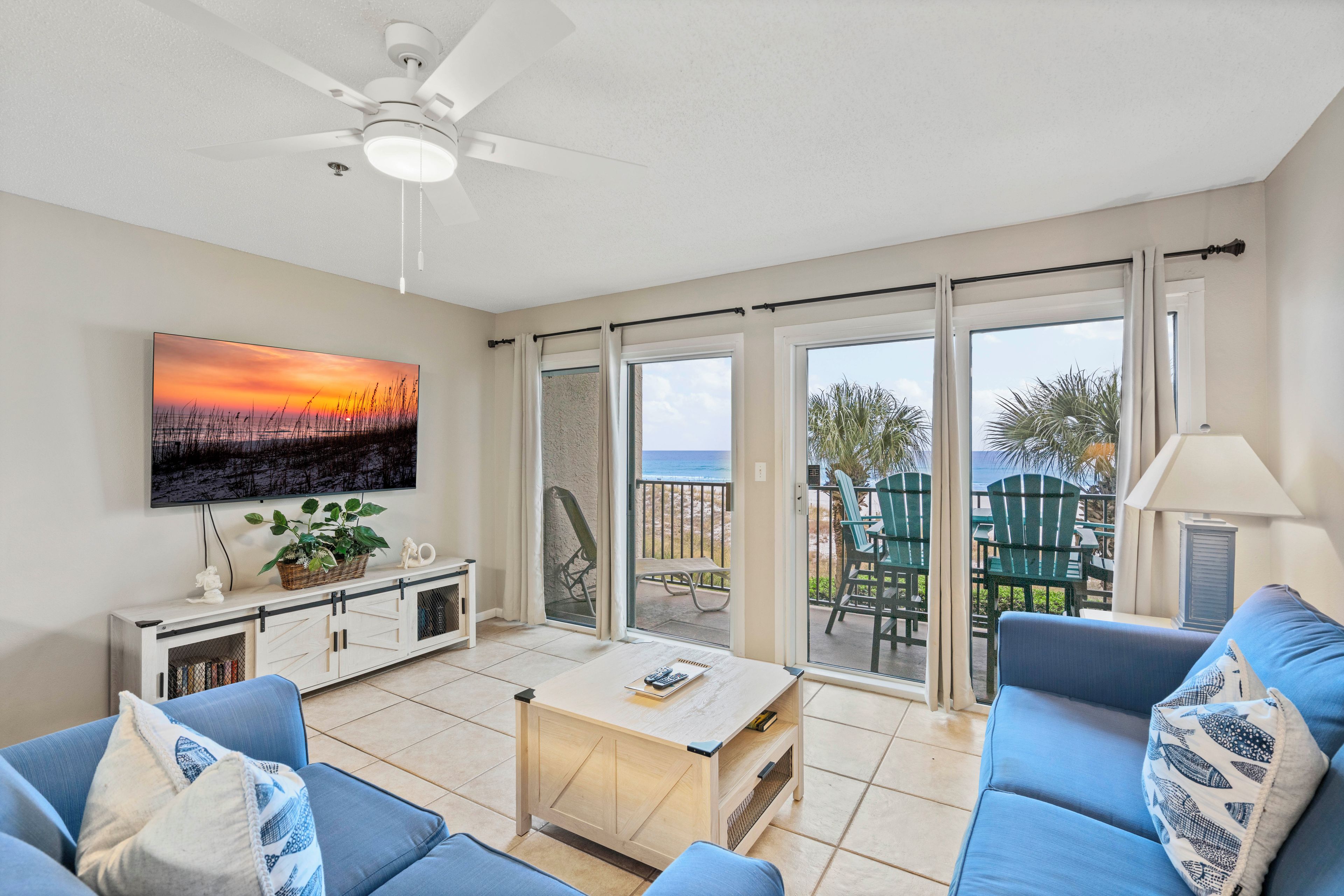 Un salon comprenant un canapé, une table basse et un téléviseur fixé au mur. Il y a un balcon avec des chaises et une vue sur la plage.