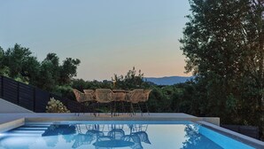 Piscina com reflexo nítido de cadeiras e árvores, cercada por vegetação e vista para colinas ao longe.