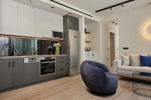 A modern kitchen with built-in appliances, a grey sofa, and a dining area with a metal chair. - Althea Suite - A Lavish Seaview Suite, suite (ZAKYNTHOS)