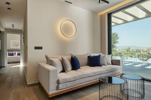 A modern living room with a sofa, a circular wall light, and a view of the outdoors. - Althea Suite - A Lavish Seaview Suite, suite (ZAKYNTHOS)
