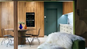 Una cocina moderna con armarios de madera, horno empotrado, comedor con mesa y sillas redondas, y una butaca blanca y mullida al frente.