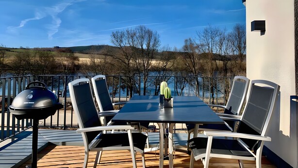 An outdoor dining area with a table and chairs, a grill, and a view of the lake and hills. - Holiday Home Mena at Lake Diemelsee (Diemelsee)