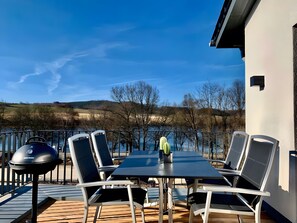 An outdoor dining area with a table and chairs, a grill, and a view of the lake and hills. - Holiday Home Mena at Lake Diemelsee (Diemelsee)