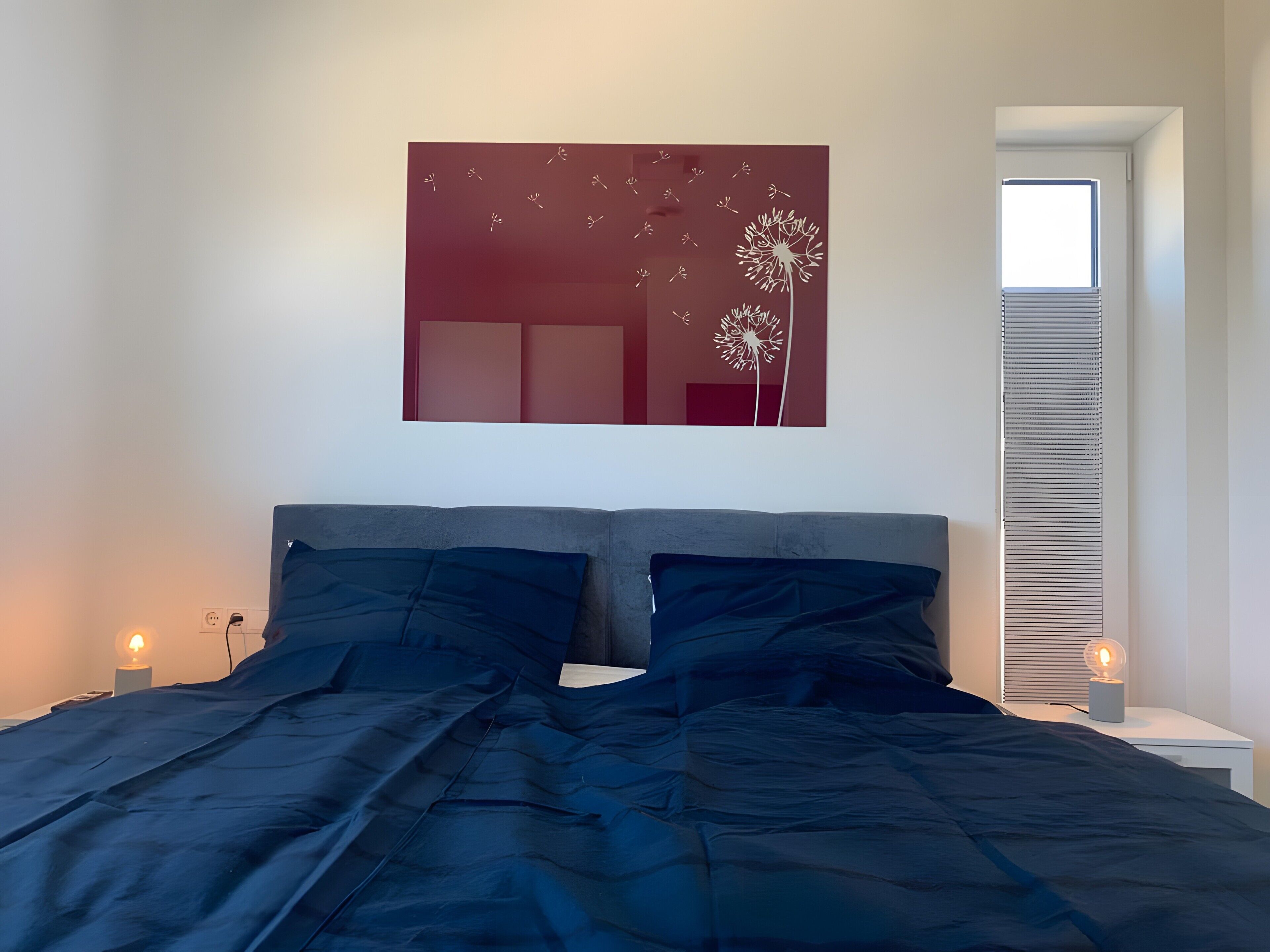A bedroom with a bed, a wall-mounted artwork, a window, and a bedside table with a lamp.