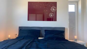 A bedroom with a bed, a wall-mounted artwork, a window, and a bedside table with a lamp.