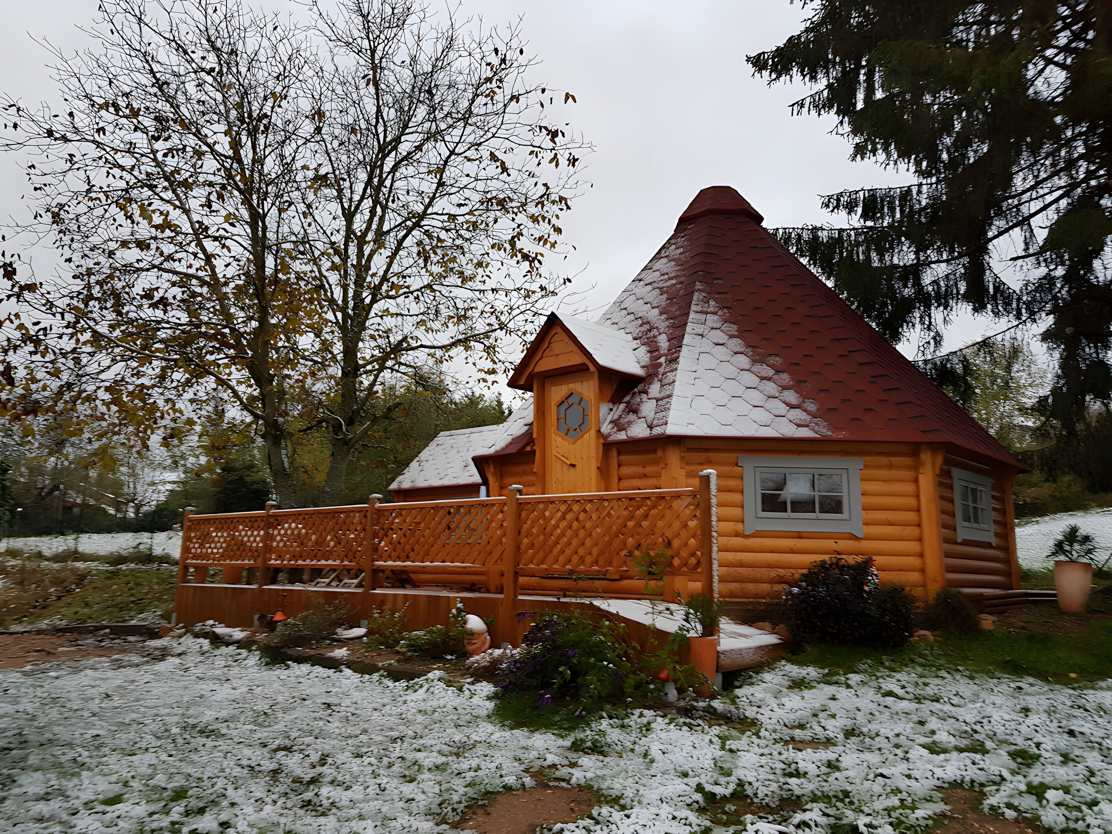 A log cabin with a snow-covered roof, a wooden fence, and a garden with snow.