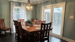 A dining area with a wooden table and chairs, a chandelier, and a view through a window with blinds. - Old Village Home w/ Huge Patio-Beach & CHS in 10 Mins! STR#ST260017 BL#20124322 (Mount Pleasant)