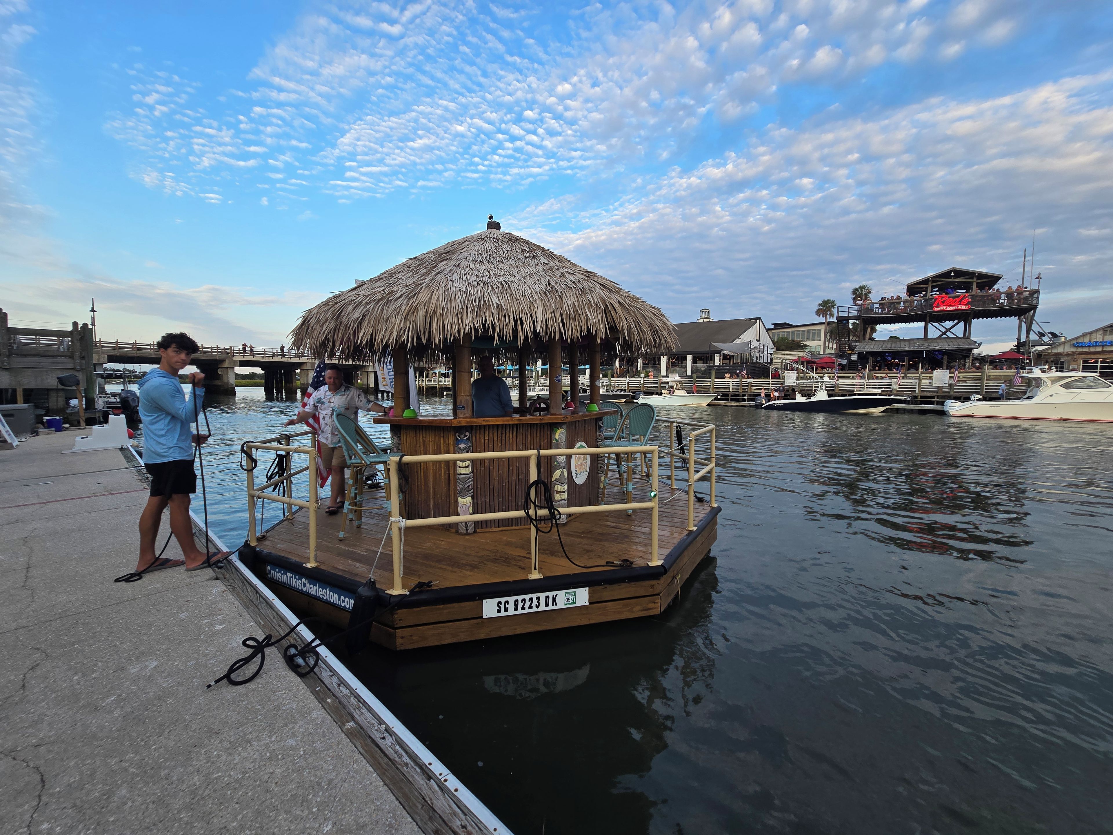 A docked boat with a thatched roof, a wooden deck, and a small bar area.