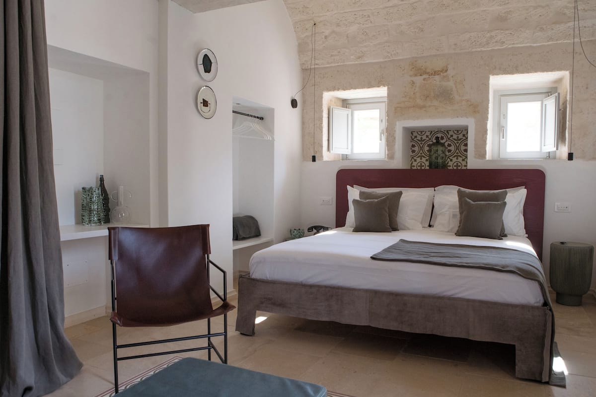 Un dormitorio con una cama grande, una silla, una mesita y una ventana con cortinas.