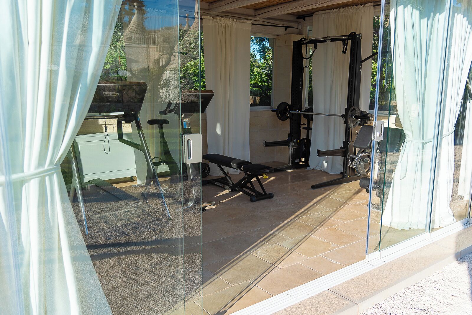 A home gym with exercise equipment, including a bench press and a squat rack, in a sunlit room with large windows.