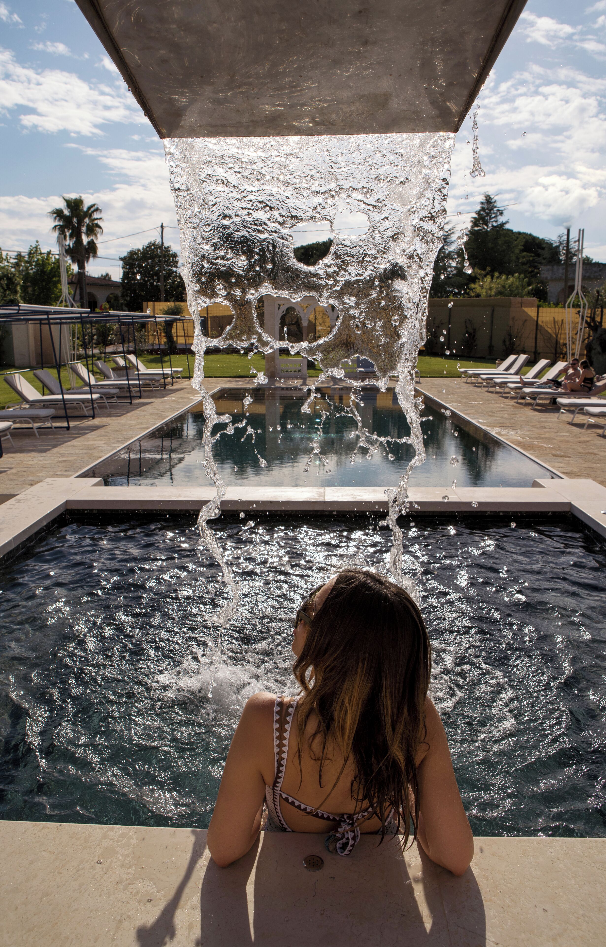 Una piscina con agua que cae desde una estructura superior, rodeada de sillas de descanso y un cielo despejado.