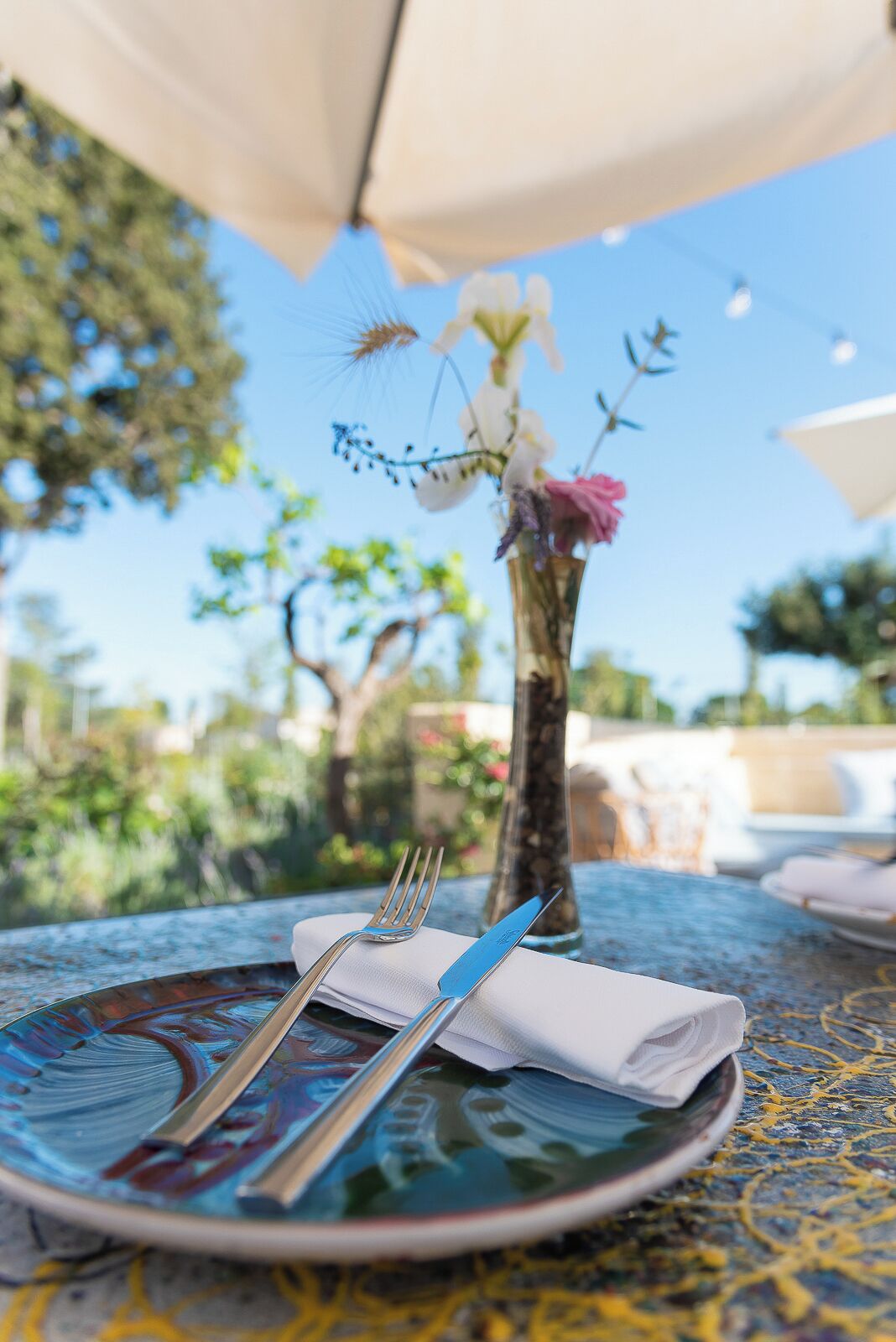 A table set outdoors with a vase of flowers, a napkin, and cutlery.