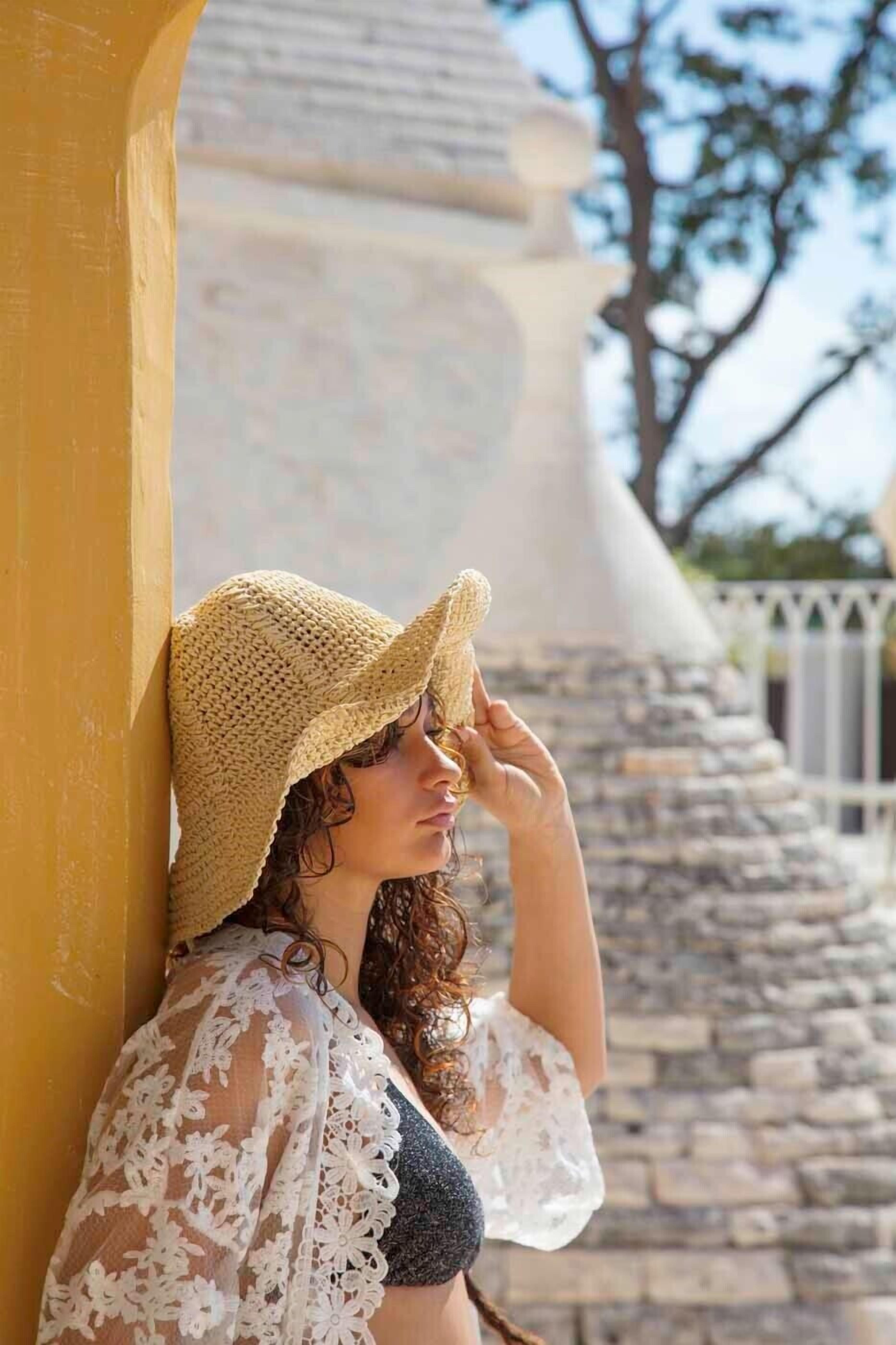 Una persona apoyada en una pared amarilla, con sombrero de paja y un vestido de encaje.