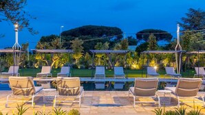 Zona junto a la piscina con sillones de descanso y una pérgola, rodeada de vegetación y vistas a una colina.