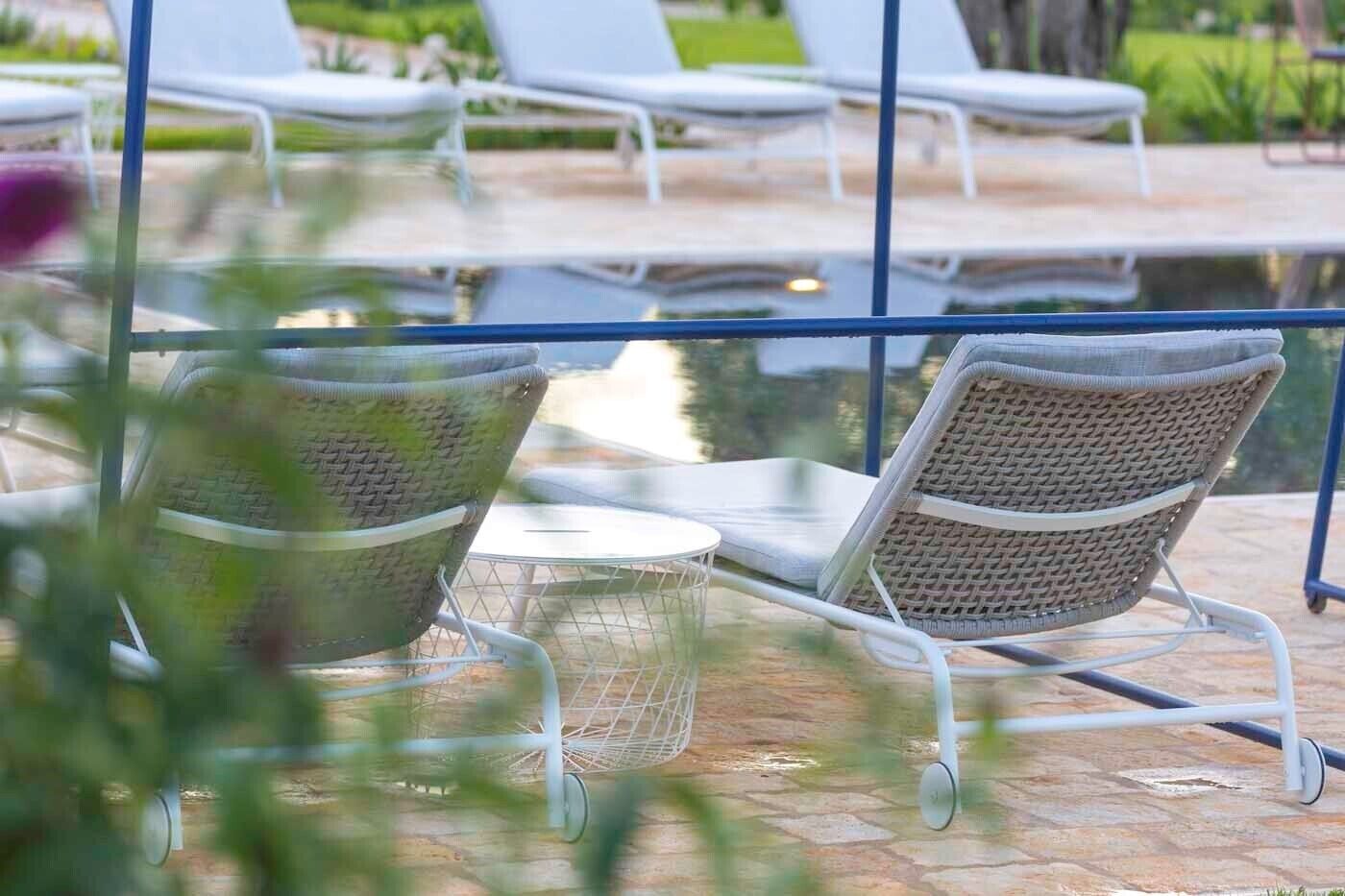 Un área junto a la piscina con sillones de descanso y una mesa, rodeada por una barandilla de vidrio.