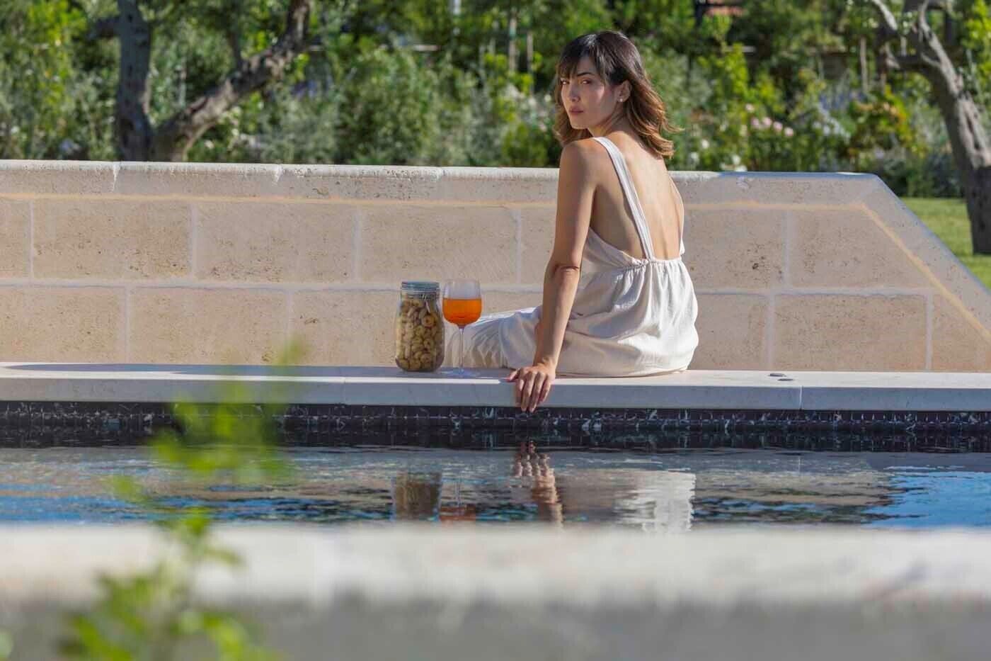 Una persona sentada junto a una piscina, con una bebida en un vaso y un tarro de frutos secos.
