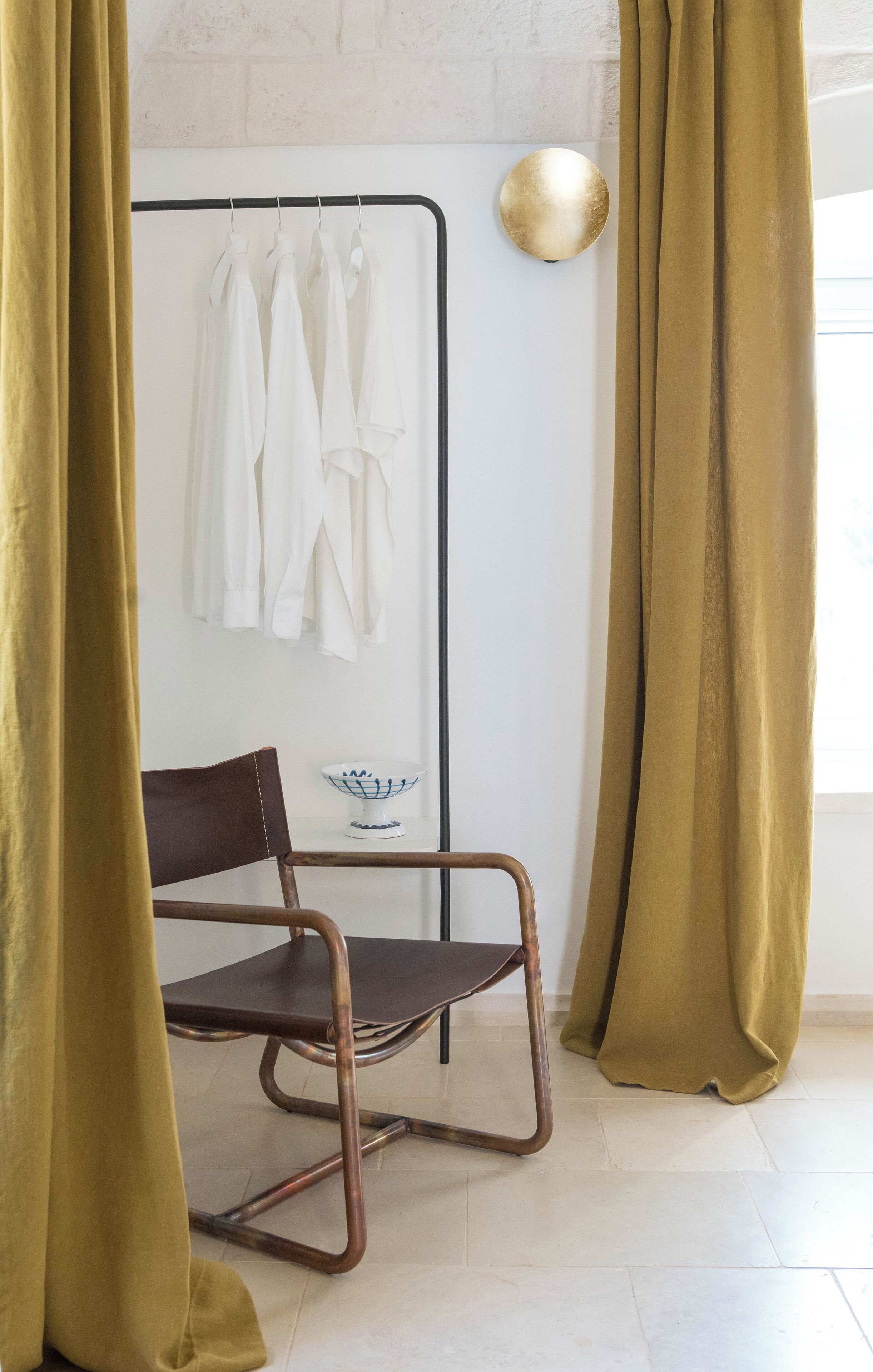 Ein Zimmer mit einem Sessel, einem Kleiderhaken mit weißen Hemden, einer Schale auf einem Tisch und einer Wandleuchte.