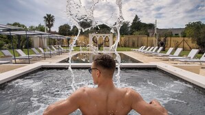 Un hombre se relaja en una piscina con salpicaduras de agua, rodeado de tumbonas y una pérgola.
