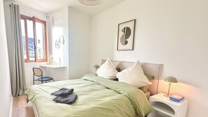 A bedroom with a bed, a nightstand, a lamp, a chair, a window with curtains, and a picture on the wall. - Direct metro to central Paris 3-rooms apartment with Panoramic View-Free Park (Saint-Denis)