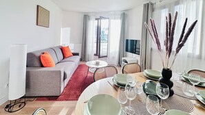 A modern living room with a grey sofa, red cushions, a dining table set with green plates and wine glasses, and a large window with curtains. - Direct metro to central Paris 3-rooms apartment with Panoramic View-Free Park (Saint-Denis)