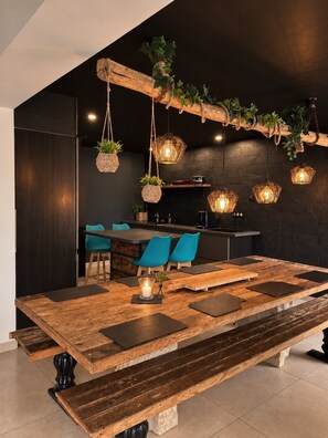Un comedor moderno con una mesa de madera, sillas azules, lámparas de mimbre colgantes y una pared de ladrillo negro.