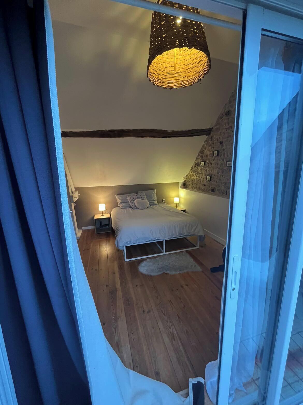 Un dormitorio con techo inclinado, una cama con almohadas, lámparas de noche y una luminaria tejida.